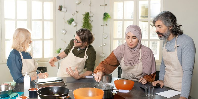 Young Muslim female trainee in hijab holding whisker over pan with water on electric stove while cooking at masterclass among other learners